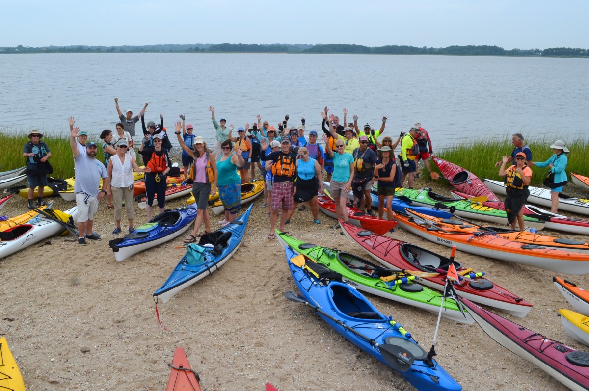 30 people in colorful gear wave on a beach with kayaks