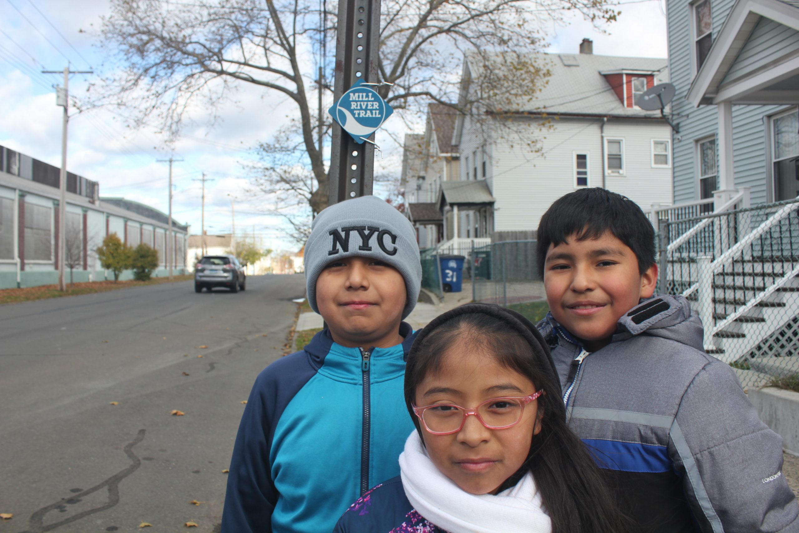 Three Latinx children in winter clothing stand on a sidewalk with a Mill River Trail marker